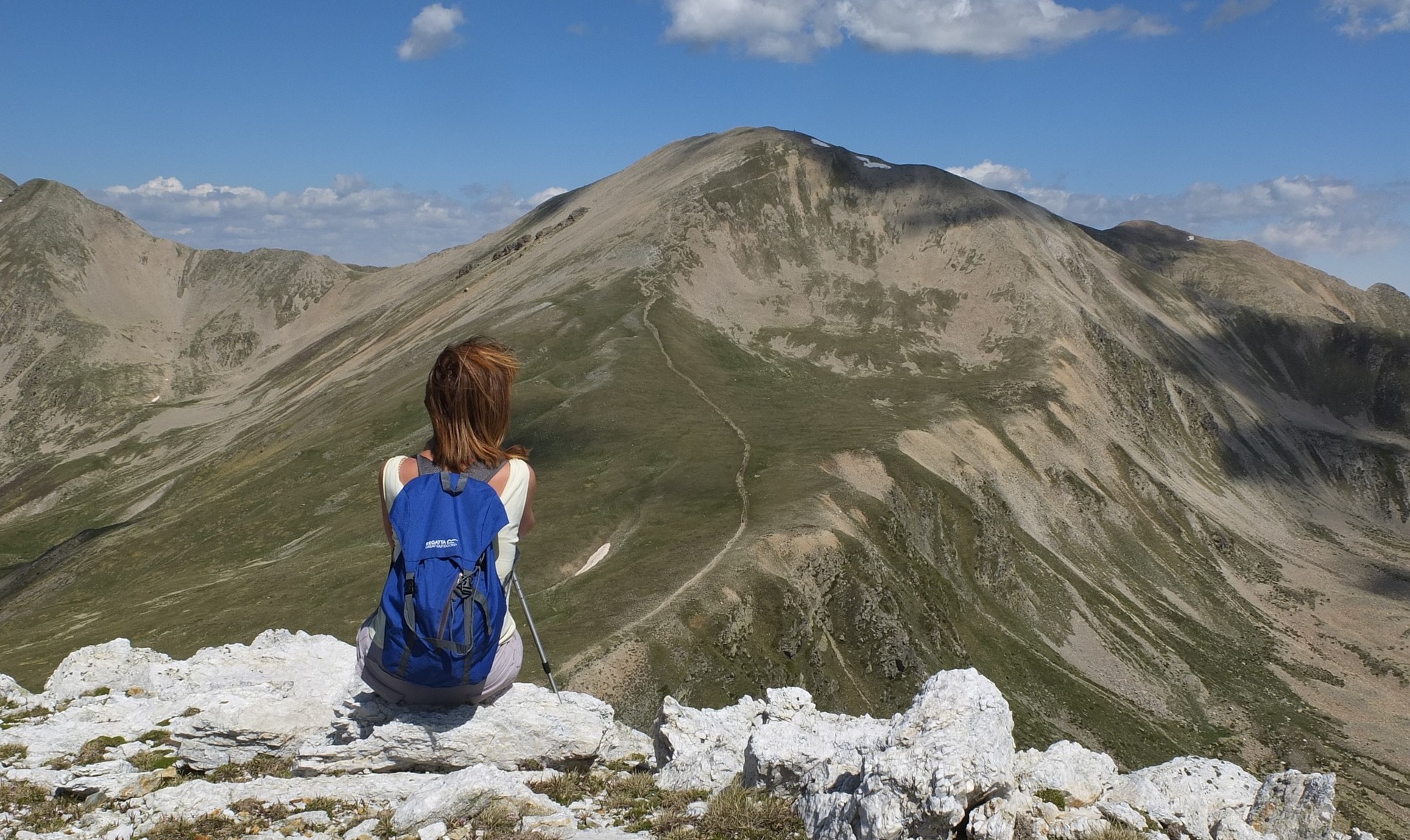 A young woman sits with her back to the camera facing a mountain. A path winds up the mountain.