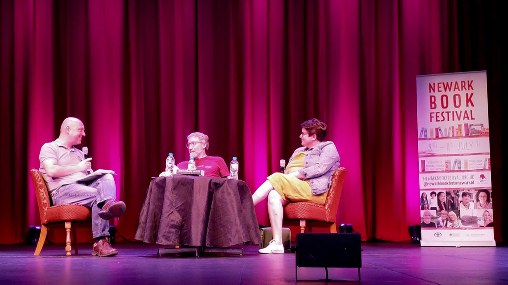 Panel at Newark Book festival. The moderator and two authors sit around a table on a stage with a purple curtain behind.