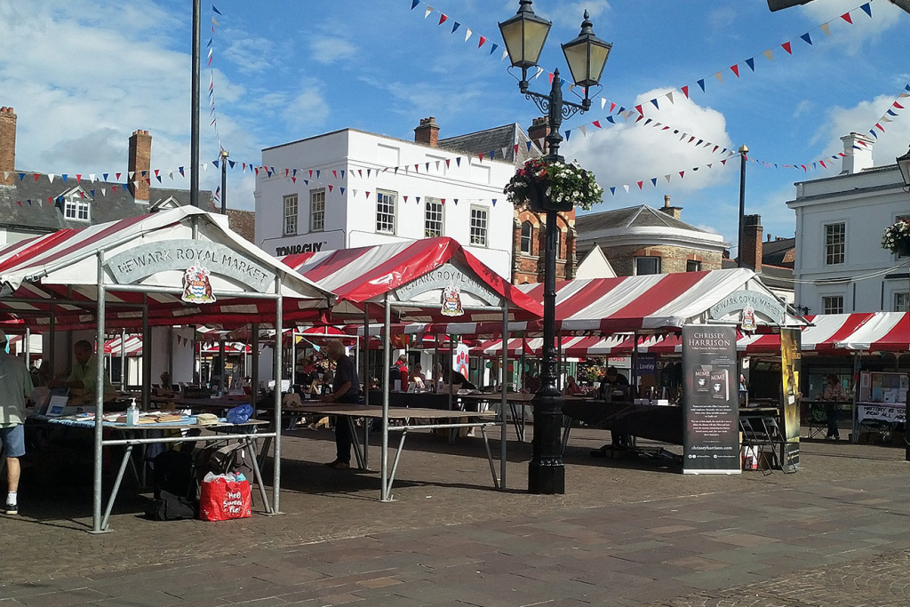 Newark-on-Trent market square. Red and white canopies of market stalls, bunting strung from a Victorian style lamp post. Blue sky.